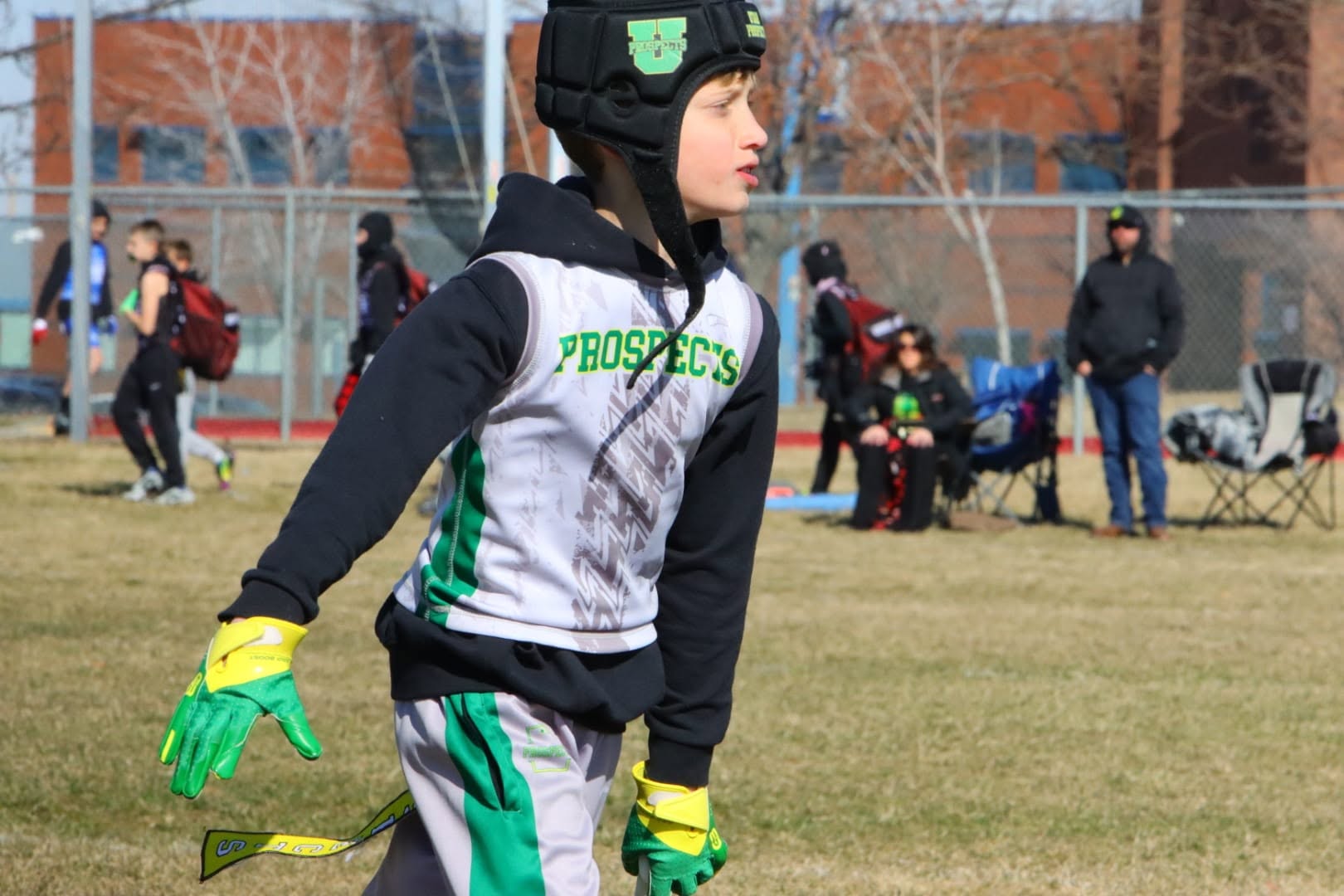 A young Utah Prospects flag player in a Prospects vest, soft-shell helmet and green-and-yellow gloves on the field